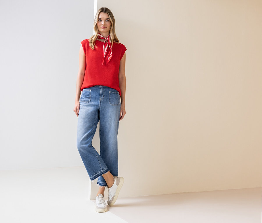 Žena stojí a má na sebe červený top, šatku na krk, džínsy a tenisky.
