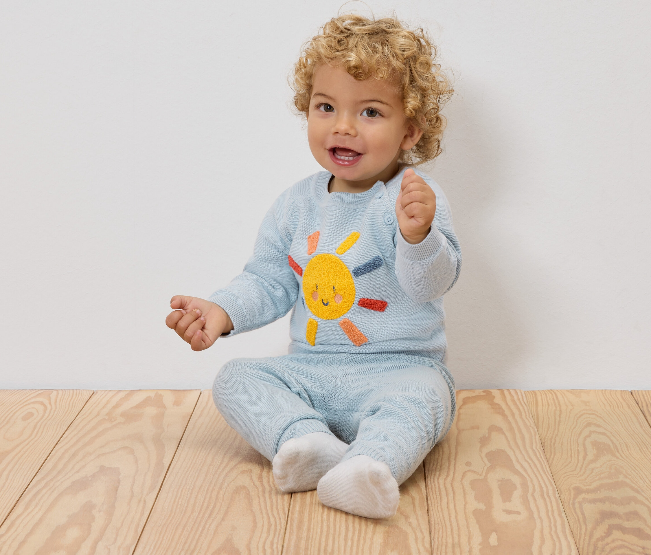 Usmievavé dieťa s blond kučerami sedí na drevenej podlahe a má na sebe svetlomodrý sveter s vyšitým slnkom a zodpovedajúce nohavice.