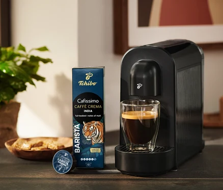 Čierny kávovar Tchibo Cafissimo so šálkou kávy, vedľa balenie kávových kapsúl Tchibo Cafissimo Caffè Crema India a jedna kapsula.