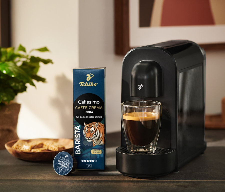 Čierny kávovar Tchibo Cafissimo so šálkou kávy, vedľa balenie kávových kapsúl Tchibo Cafissimo Caffè Crema India a jedna kapsula.