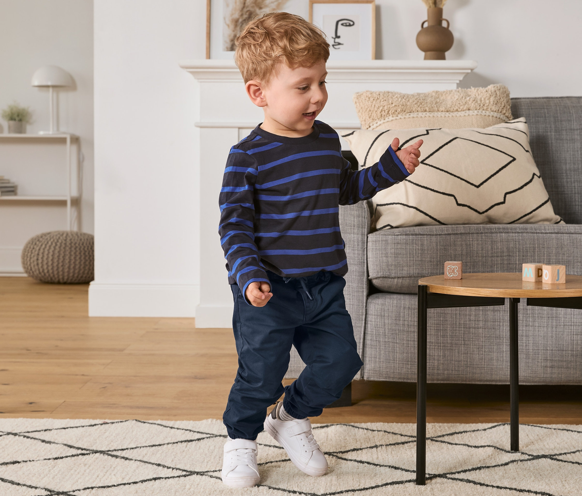 Chlapček sa hrá s kockami na stolíku, má na sebe modré nohavice chino s pružným pásom a pruhované tričko.