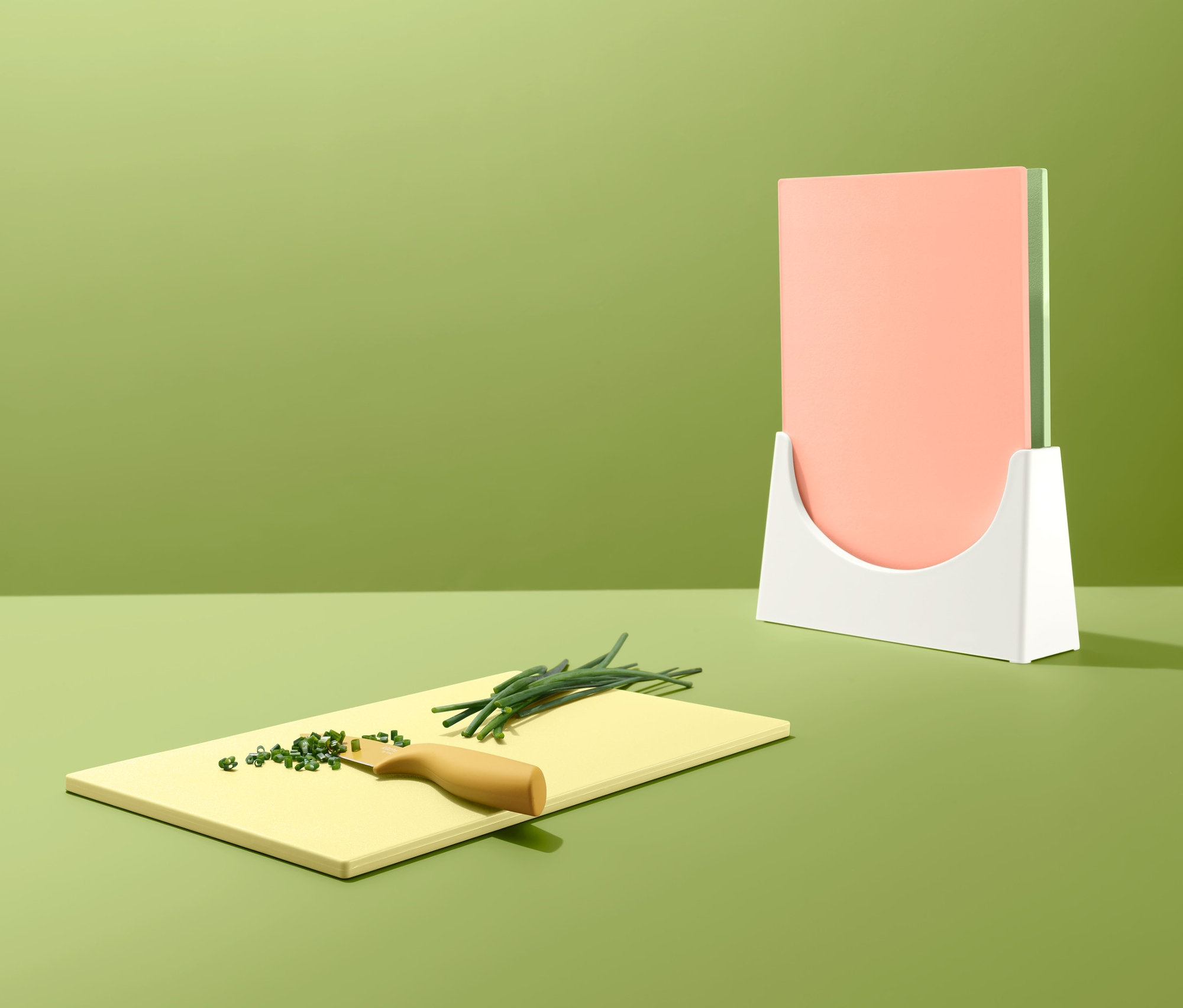 Žltá doska na krájanie so zelenou pažítkou a nožom. V pozadí stoja v držiaku ďalšie dve dosky na krájanie.