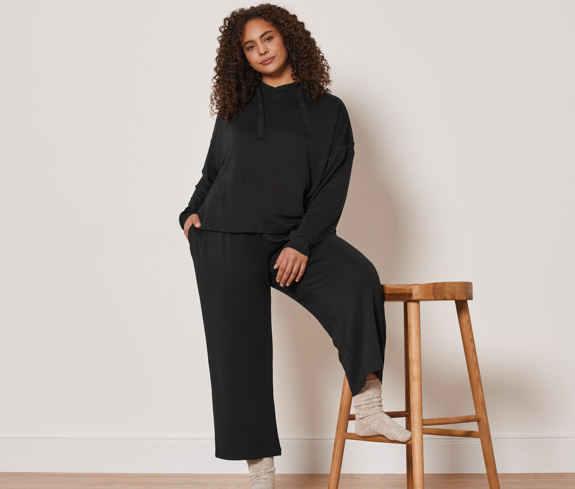 Žena pózuje opretá o drevenú stoličku a má na sebe čiernu sedemosminovú domácu súpravu.