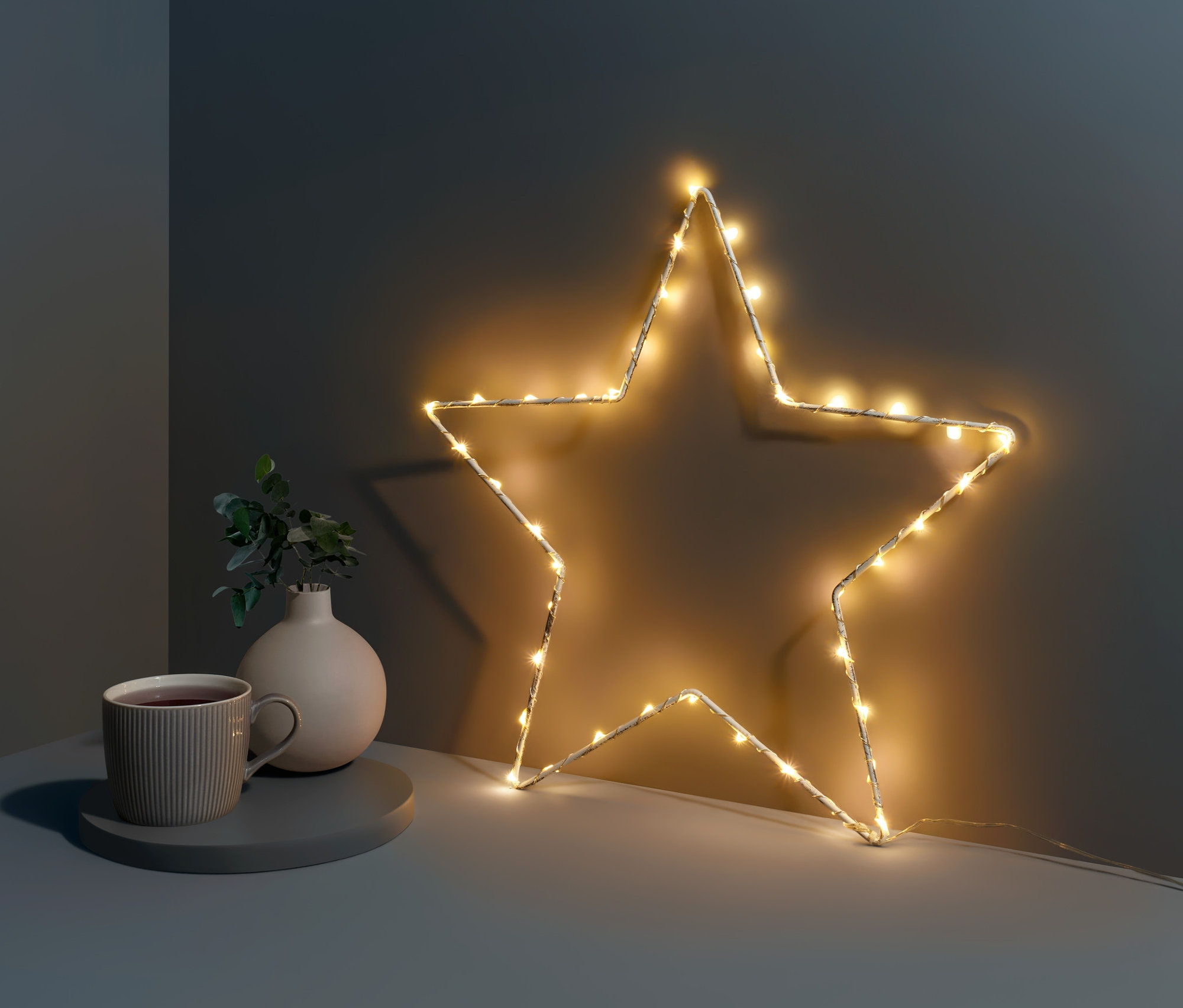 Malá kovová hviezda s LED diódami stojí na stole vedľa šálky a vázy.