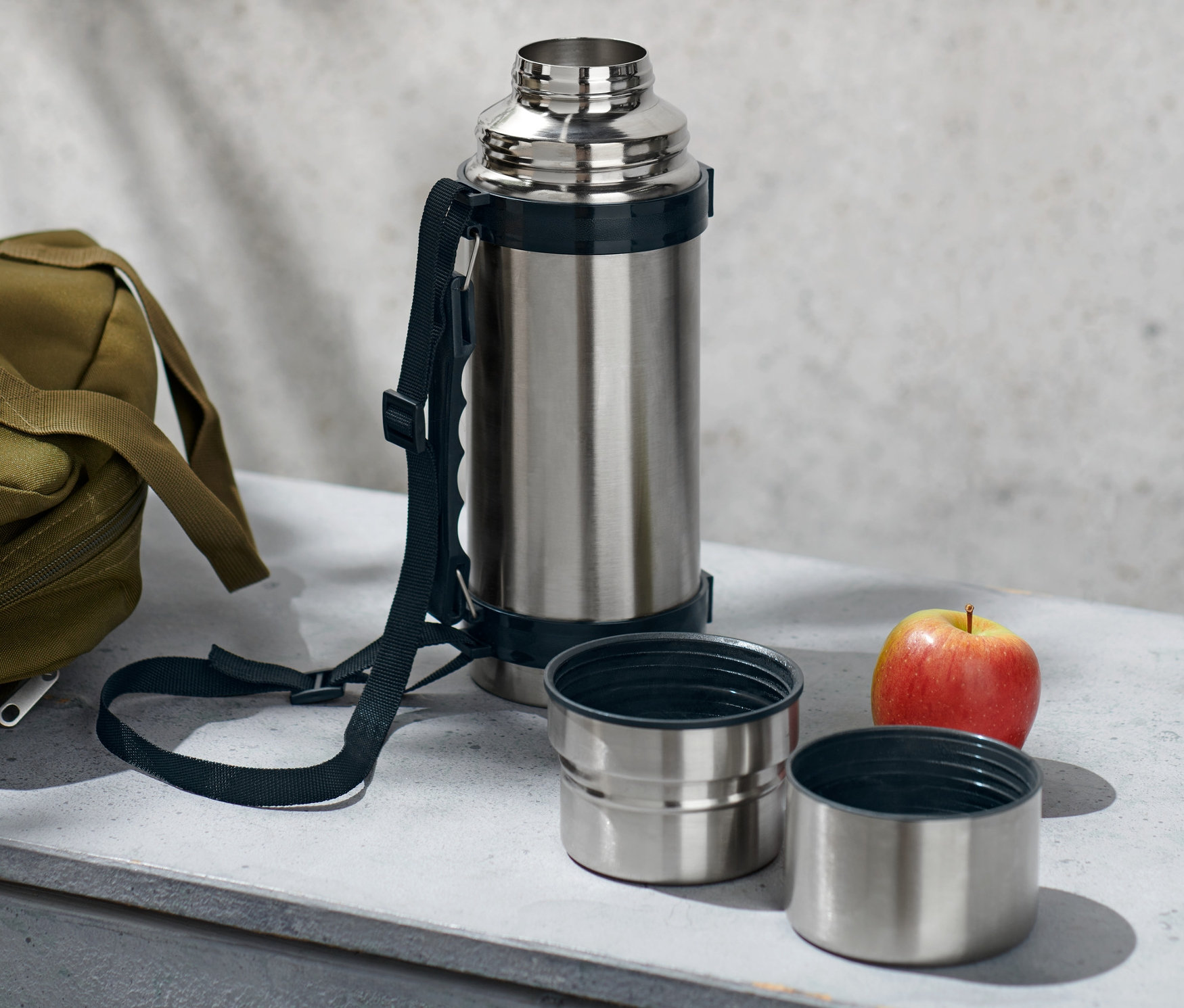 Strieborná termoska s dvoma pohárikmi, olivová taška a jablko ležia na sivej ploche.