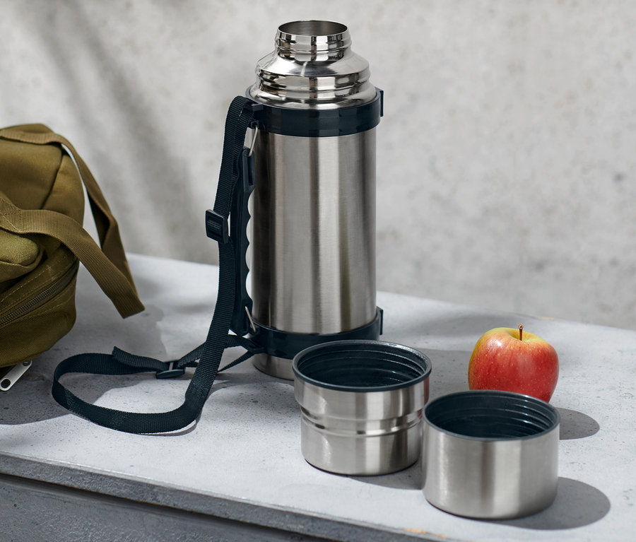 Strieborná termoska s dvoma pohárikmi, olivová taška a jablko ležia na sivej ploche.