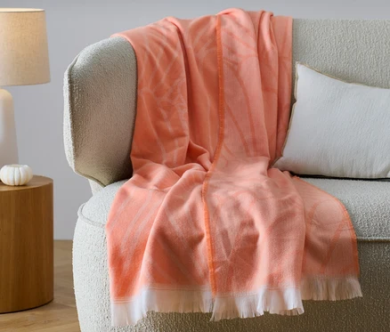 Oranžová obojstranná deka so strapcami leží na béžovej pohovke vedľa bieleho vankúša.