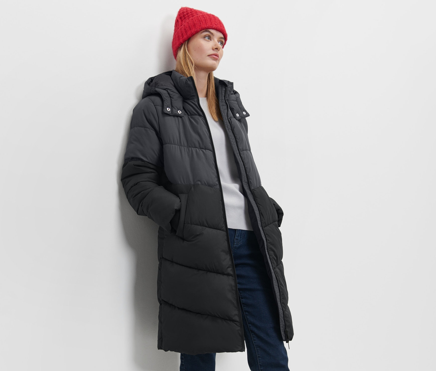 Žena pózuje v prešívanom kabáte s kapucňou a červenej čiapke.