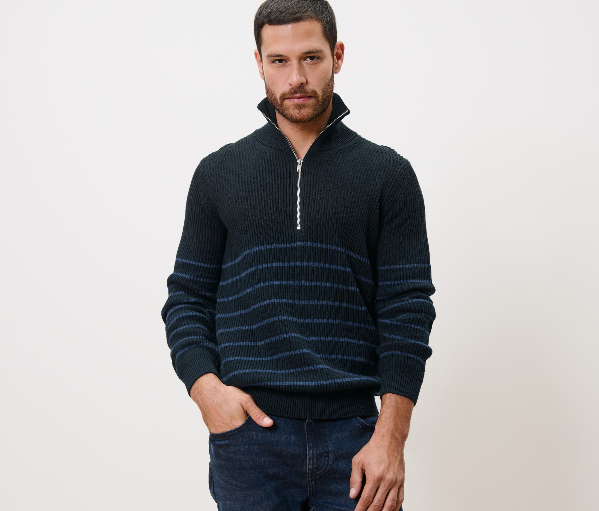 Muž pózuje v pulóvri typu troyer s rukou vo vrecku džínsov.
