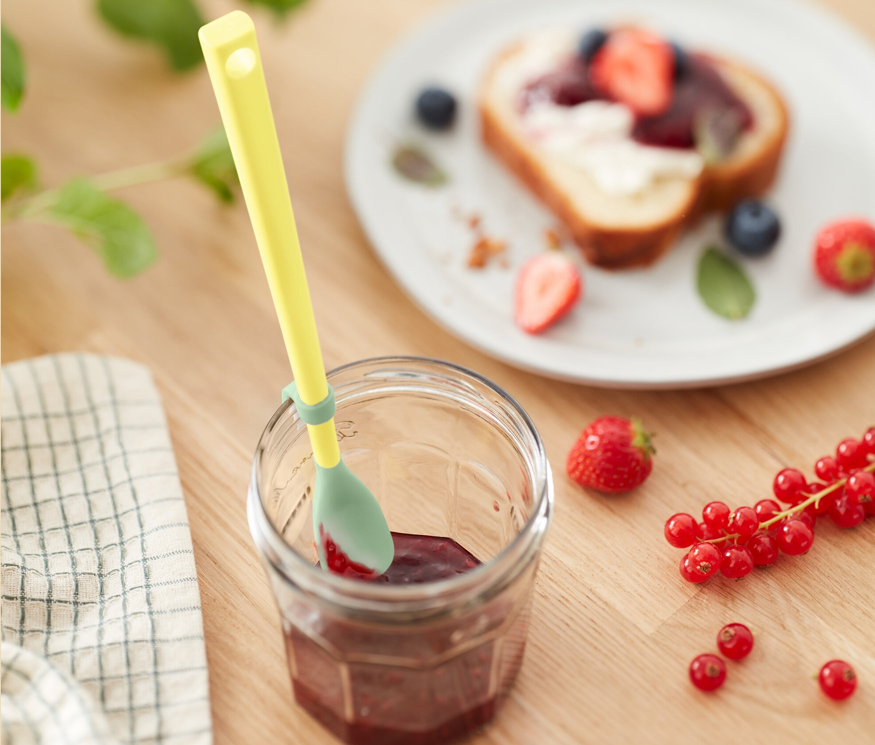 Žltá silikónová lyžica v pohári džemu vedľa taniera s toastom s ovocím.