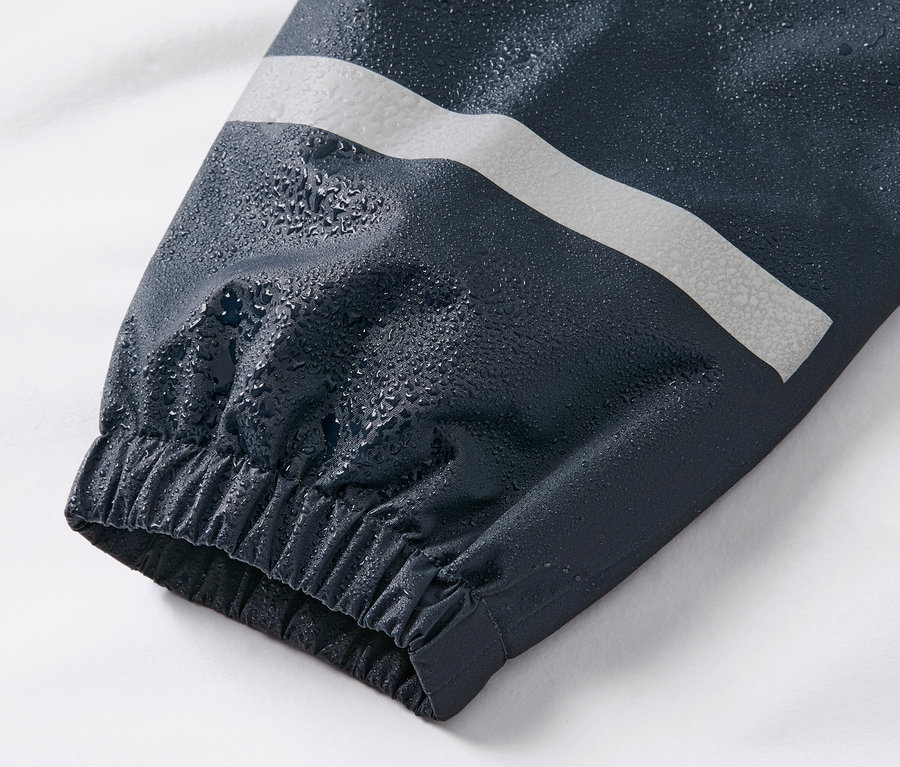 Detailný záber na rukáv detských nohavíc do dažďa tmavomodrej farby s reflexným pruhom a kvapkami vody.