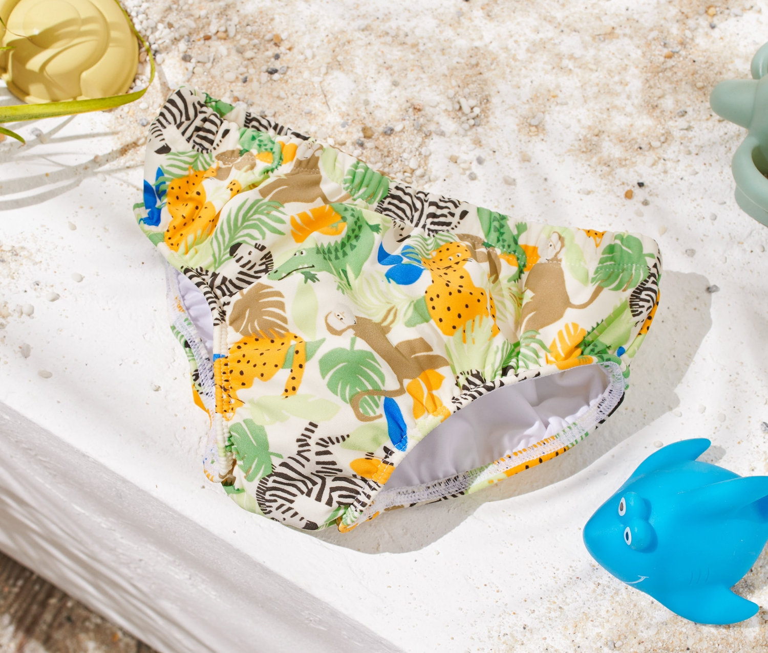 Detská plienka na plávanie so zvieracou potlačou leží na bielom povrchu vedľa hračiek do piesku.
