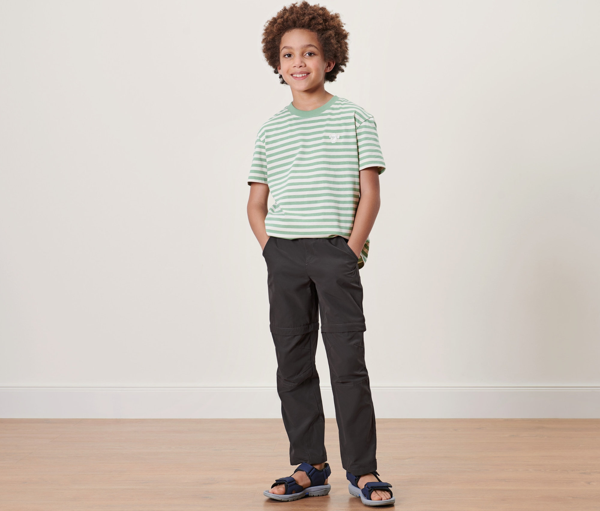 Chlapec stojí a má na sebe detské funkčné nohavice, pruhované tričko a sandále.