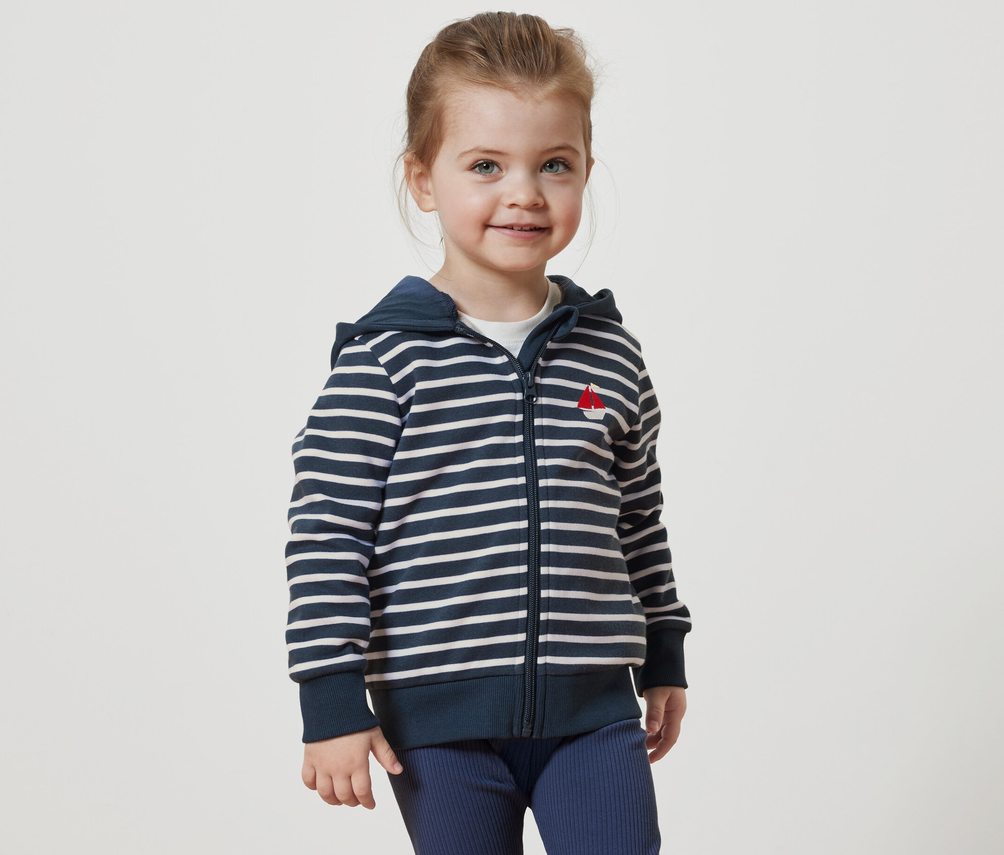 Usmievajúce sa dievčatko má na sebe pruhovanú mikinu s kapucňou a červenou výšivkou loďky.