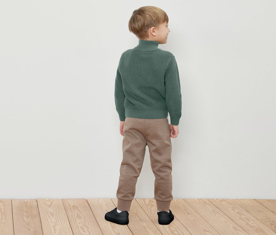 Chlapec stojí chrbtom, má na sebe zelený rolák, hnedé tepláky a čierne tenisky.