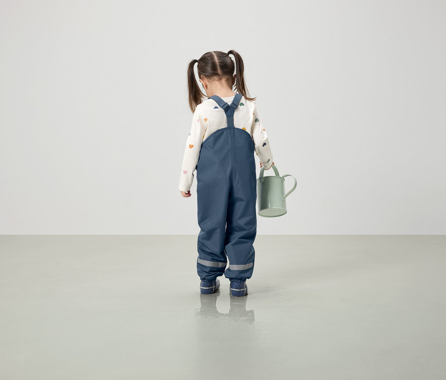 Dievča v modrých nohaviciach s trakmi drží zelenú kanvu.