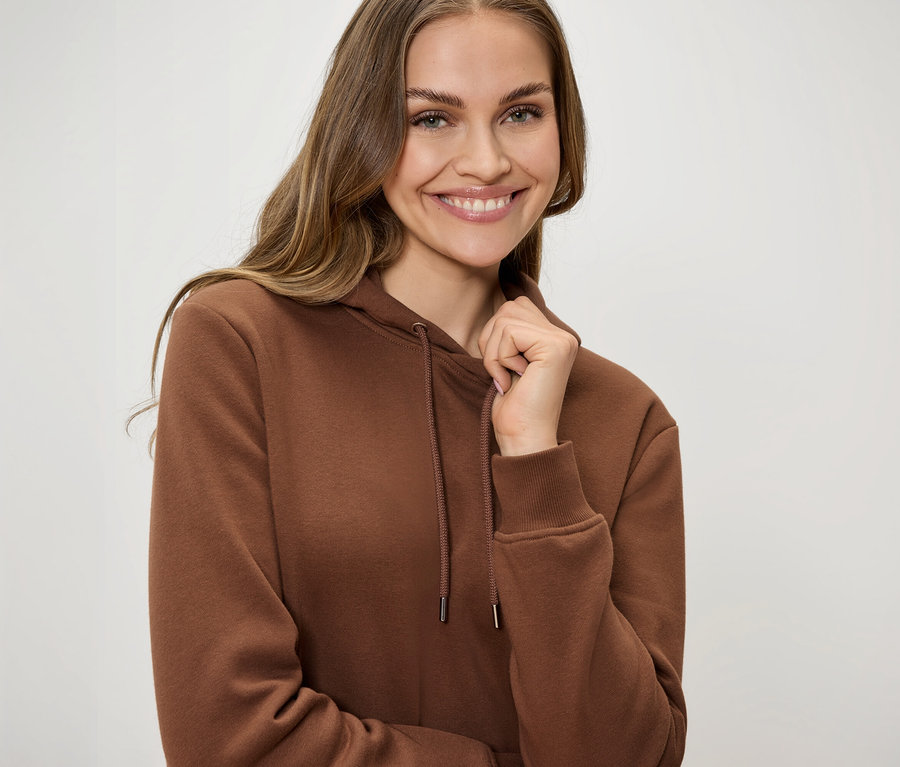 Usmievajúca sa žena v hnedej mikine s kapucňou so šnúrkami drží golier jednou rukou.