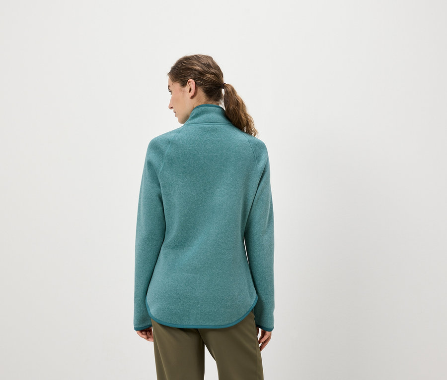 Žena chrbtom k fotoaparátu má na sebe modrozelenú flísovú bundu a olivovozelené nohavice.