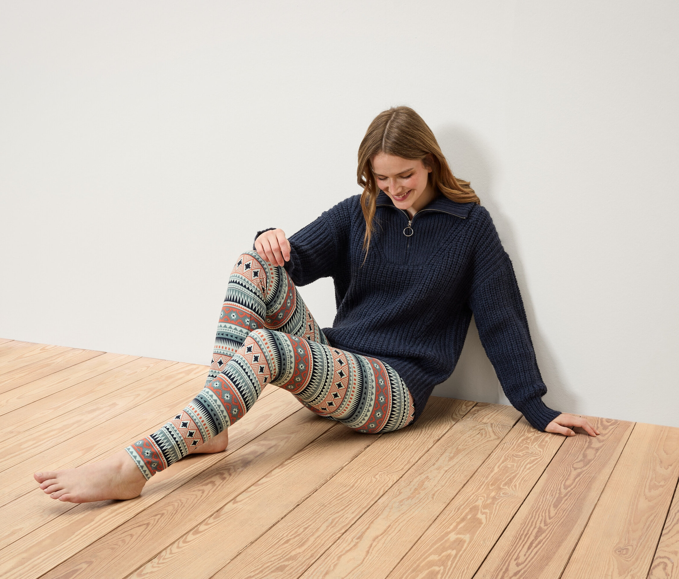 Žena sedí na drevenej podlahe, má na sebe tmavomodrý sveter na zips a vzorované legíny.