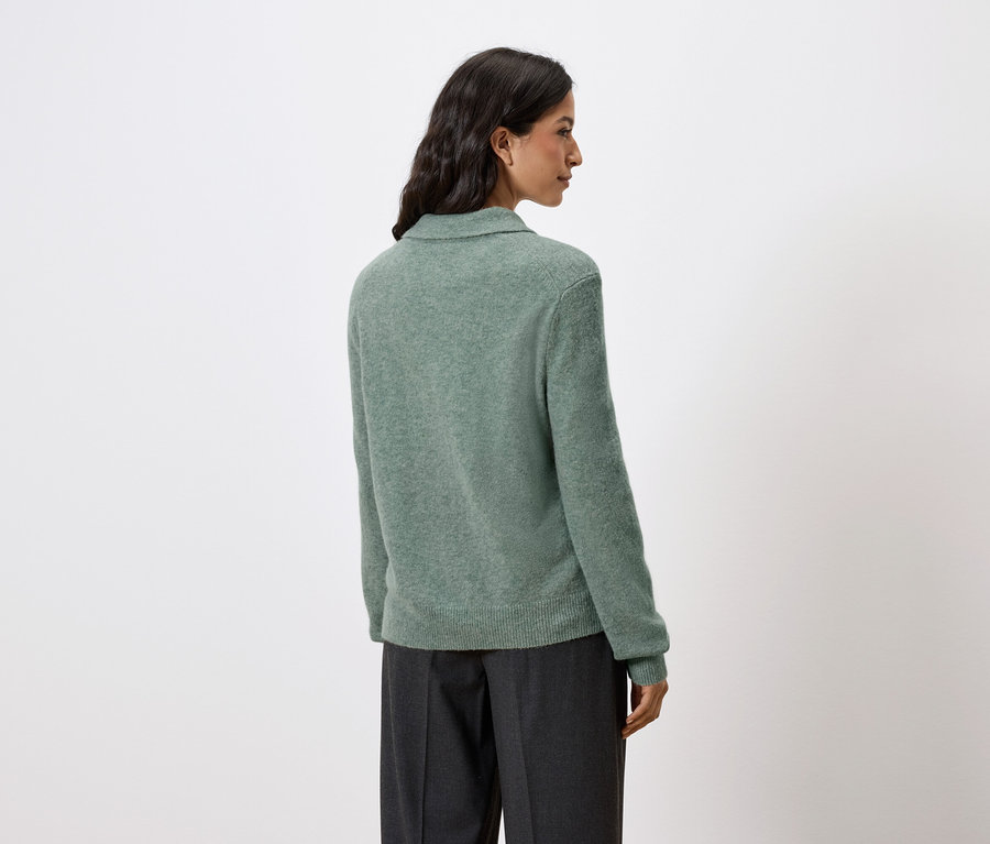 Zozadu je vidieť žena s tmavými vlasmi, ktorá má na sebe zelený svetrík a tmavé nohavice.