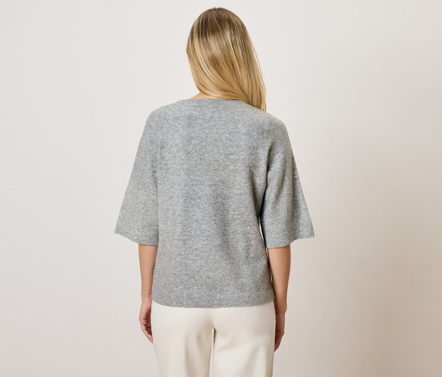 Blondínka zozadu v sivom svetri s trojštvrťovými rukávmi a bielych nohaviciach.