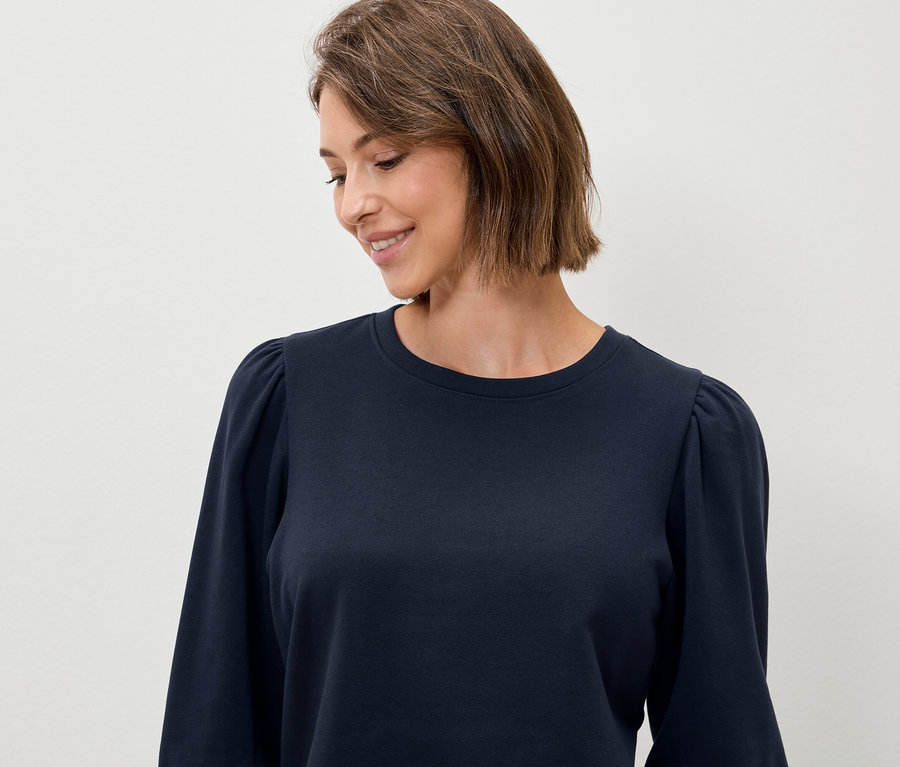 Žena s krátkymi hnedými vlasmi má na sebe tmavomodrý top s dlhými rukávmi.