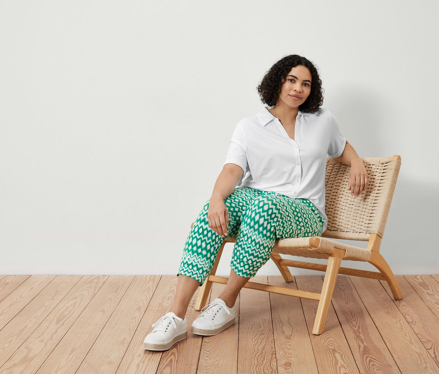 Žena sedí v kresle a má na sebe trojštvrťové džersejové nohavice s potlačou a blúzkové tričko s gombíkovou légou, biele.