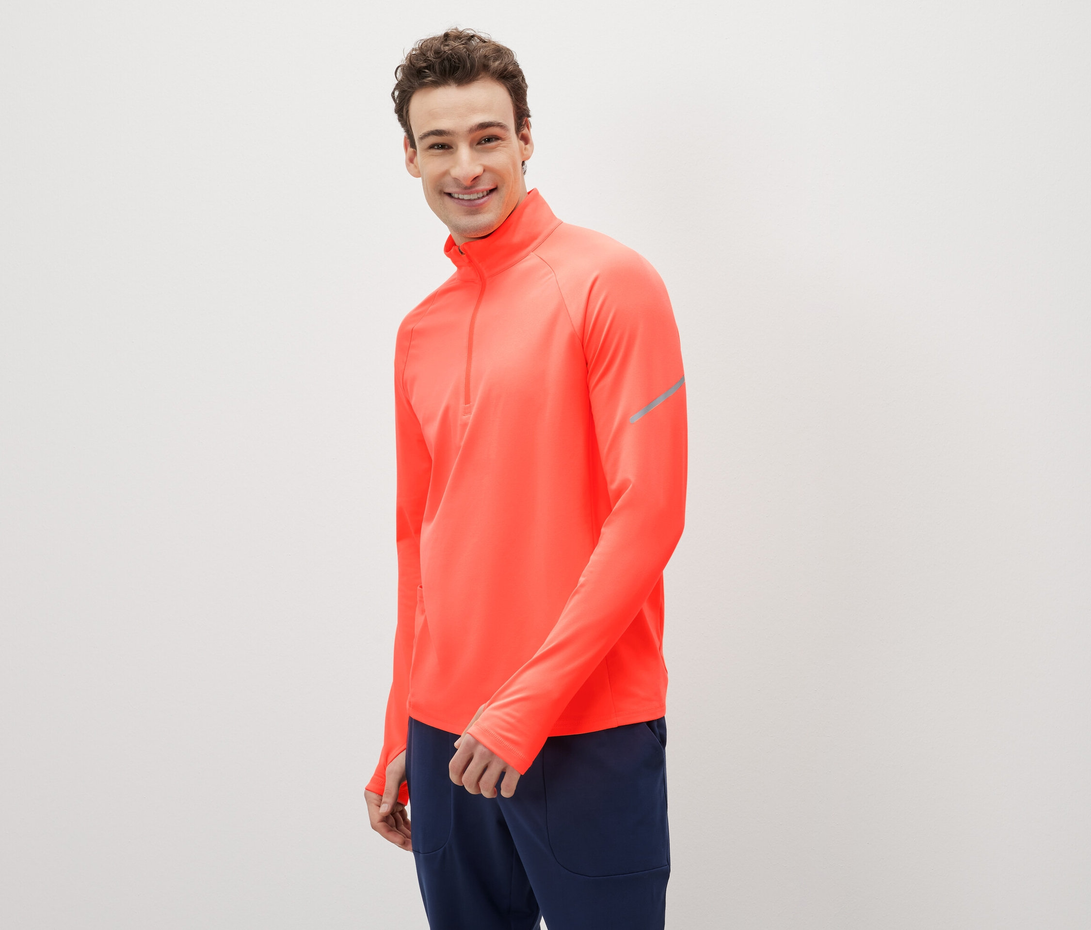 Muž sa usmieva a pózuje v koralovočervenom športovom tope s dlhým rukávom a tmavomodrých teplákoch.