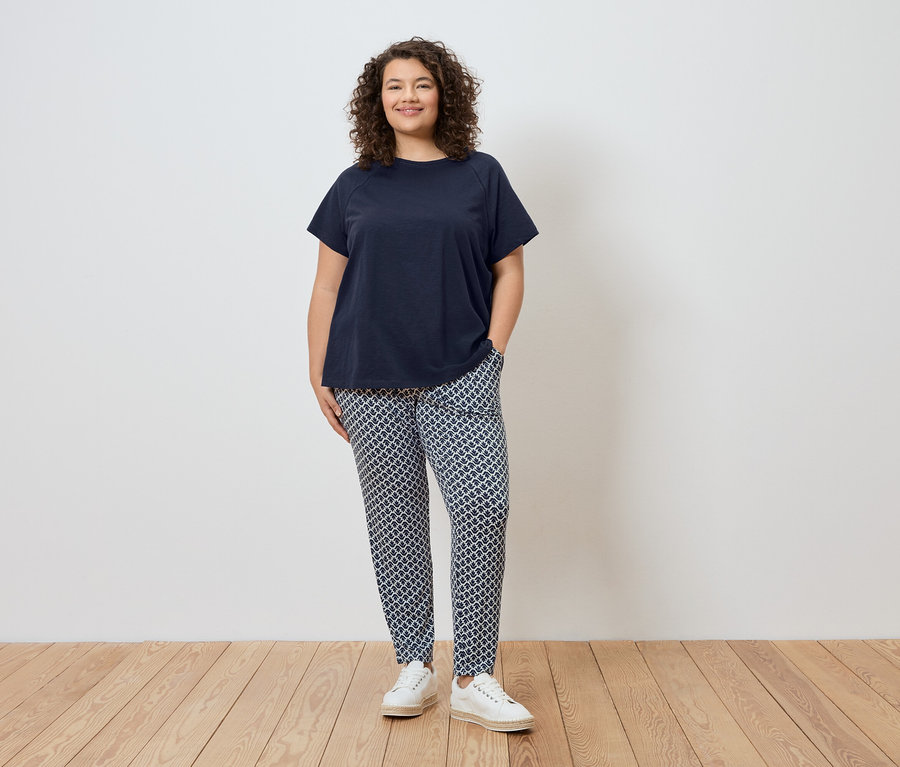 Žena stojí pred stenou, má na sebe tmavomodré tričko, vzorované nohavice a biele topánky.