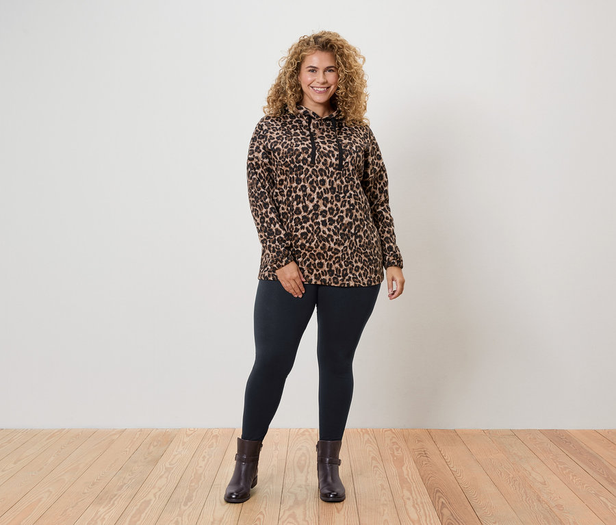 Žena s kučeravými vlasmi stojí a má na sebe top s leopardím vzorom, legíny a členkové čižmy.
