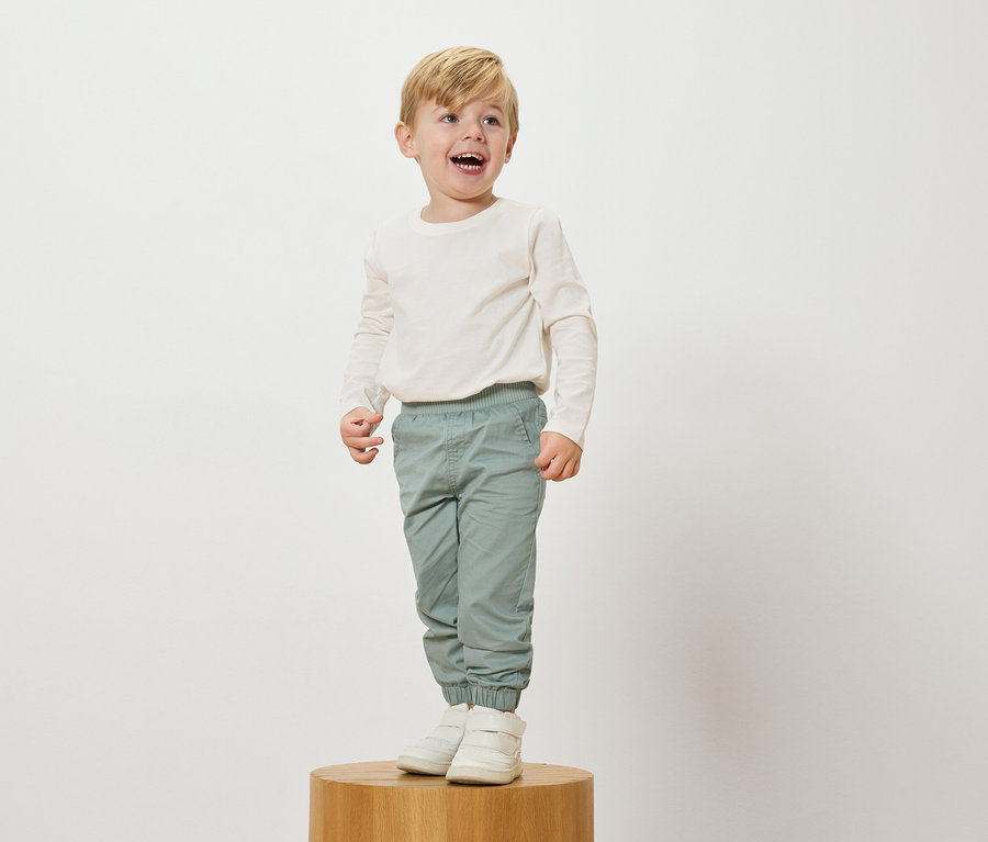 Usmievavý blond chlapec stojí na drevenom podstavci, má na sebe krémový top s dlhým rukávom, svetlomodré nohavice a biele tenisky.