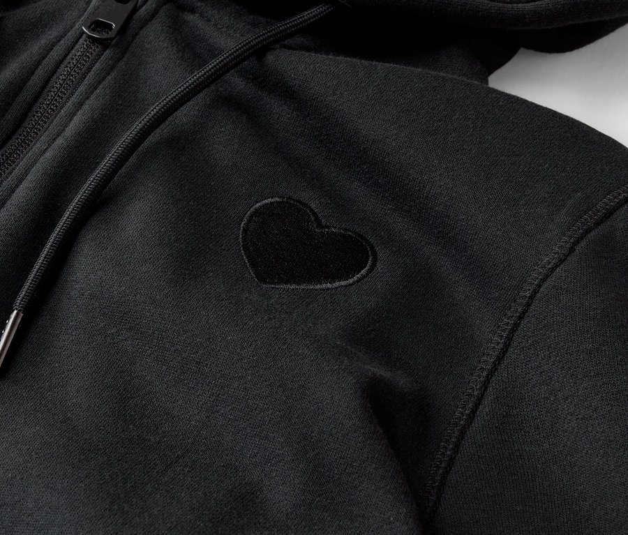 Detailný záber na čiernu mikinu s kapucňou na zips s malou aplikáciou v tvare srdca.