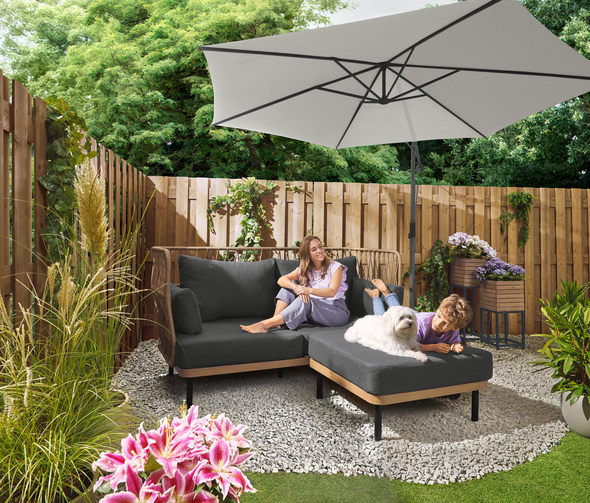 Žena sedí na sivej pohovke pod slnečníkom Leco Ampelschirm Ø 300 cm so stojanom a krytom. Dieťa leží so psom na otomane. V pozadí je vidieť sadu dosiek Leco pre krížový stojan.