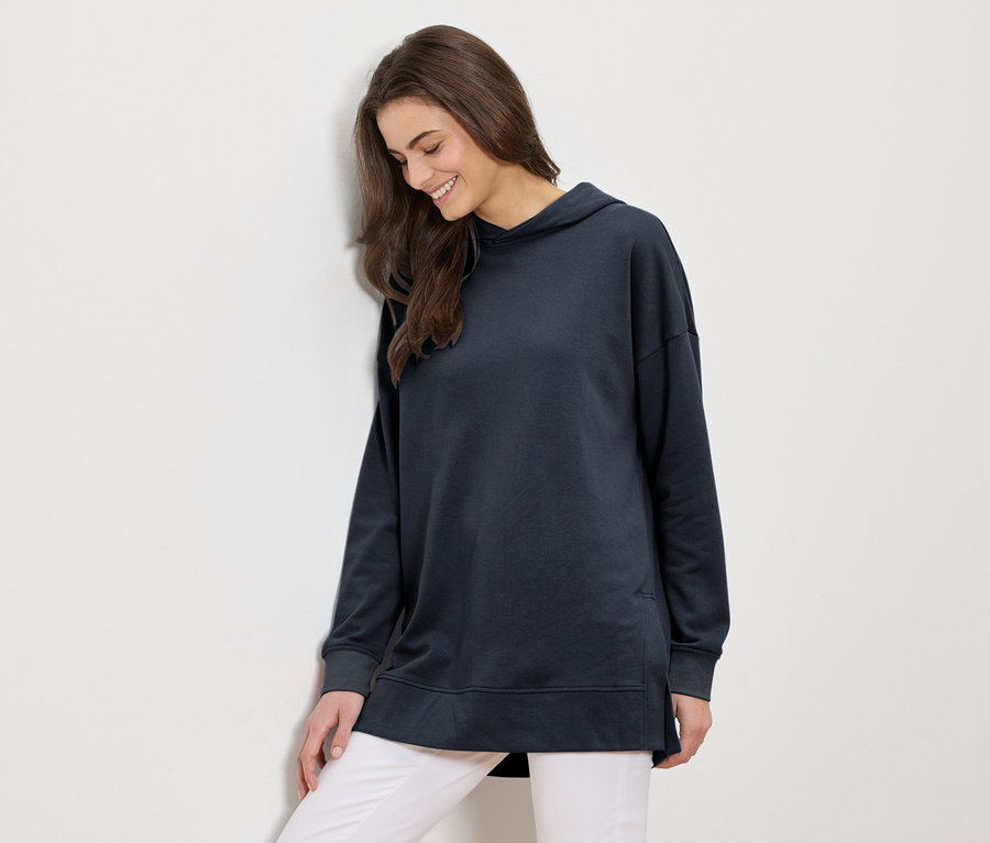 Žena s dlhými hnedými vlasmi v dlhej námorníckej modrej mikine s kapucňou a bielych nohaviciach sa usmieva a opiera sa o bielu stenu.