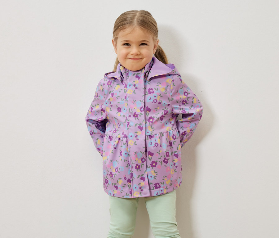 Dievča s blond vlasmi má na sebe fialovú kvetovanú bundu do dažďa a svetlozelené nohavice pred bielou stenou.