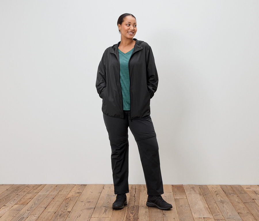Žena stojí s rukami vo vreckách v čiernej dažďovej bunde, zelenom tope a čiernych nohaviciach pred bielou stenou.
