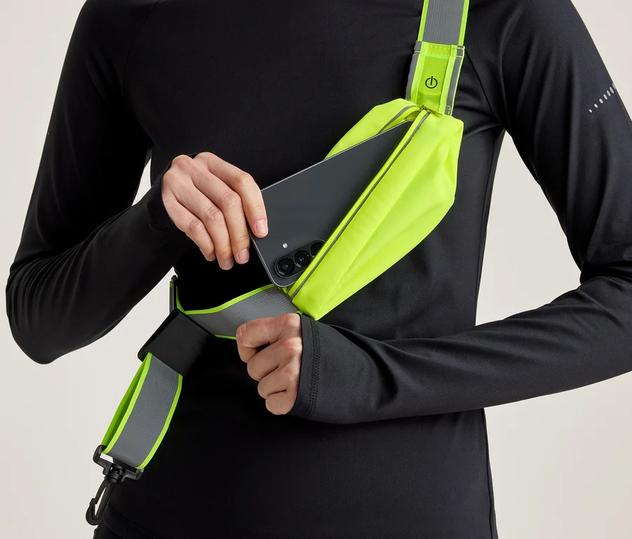 Osoba v čiernom tričku s dlhým rukávom vkladá čierny smartfón do neónovo žltej crossbody tašky s reflexným detailom.