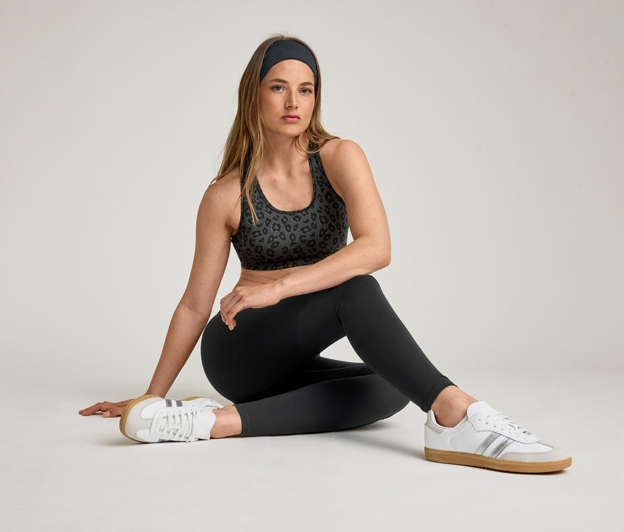 Žena sedí v športovej podprsenke, čelenke a športových legínach »ActiveSoft«. Má obuté biele tenisky.