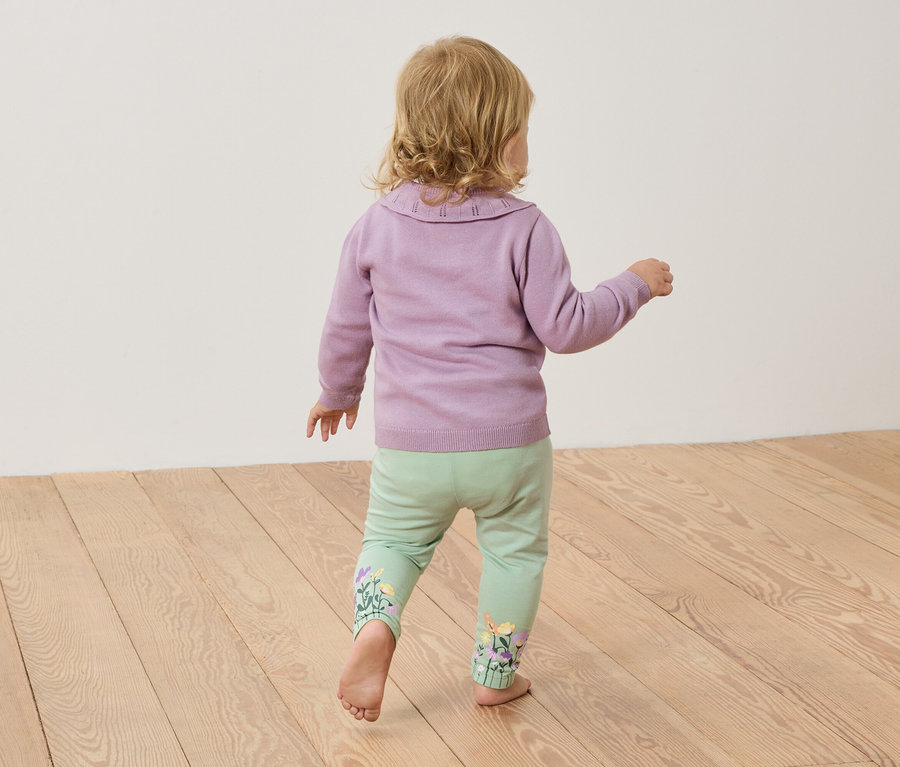 Malé dieťa s blond vlasmi stojí bosé na drevenej podlahe, má na sebe fialový sveter a svetlozelené nohavice s kvetinovou výšivkou.