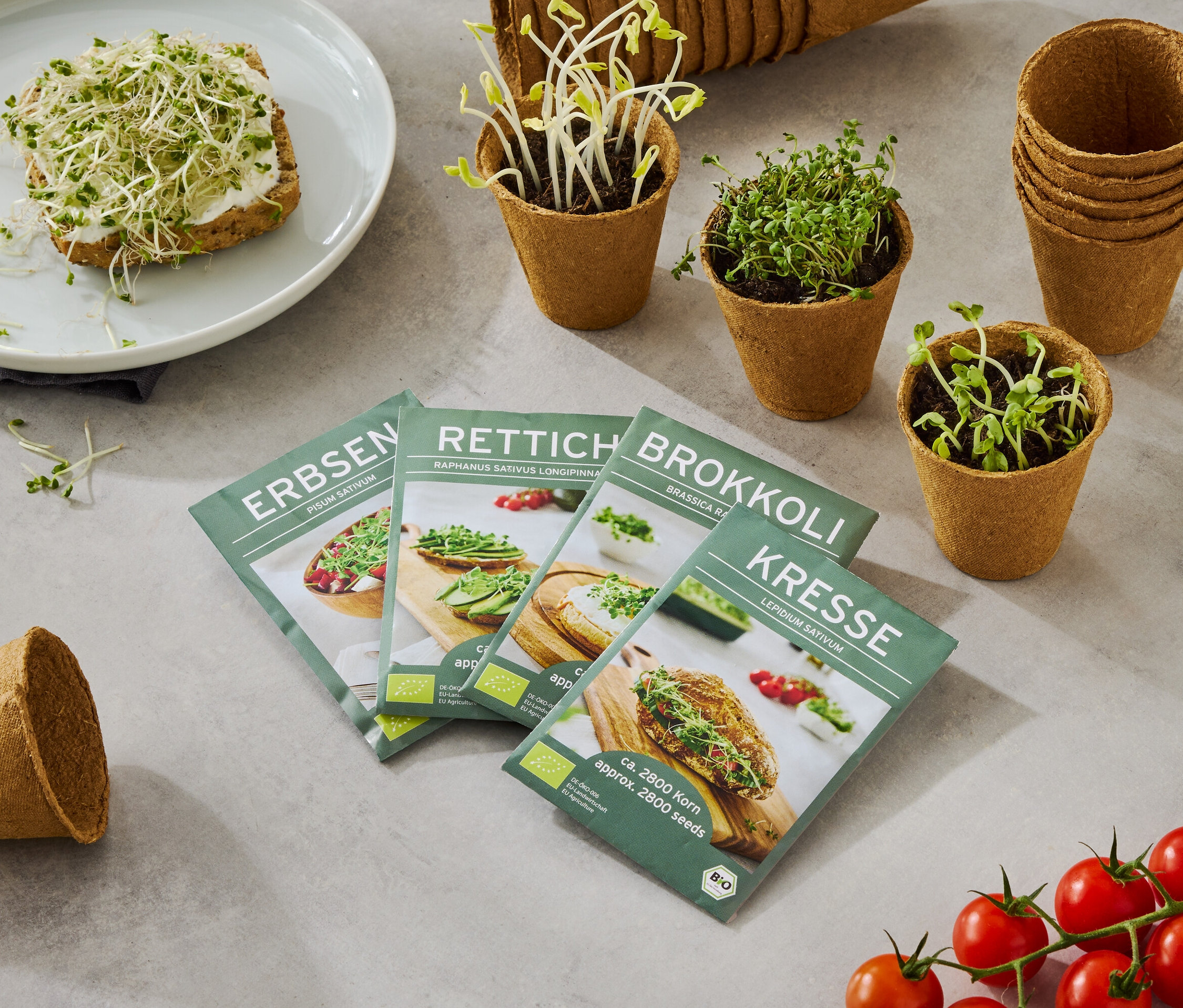 Tanier s chlebom posypaným klíčkami, spolu so štyrmi škatuľami semien (hrach, reďkovka, brokolica, žerucha), malými kvetináčmi s rastúcimi klíčkami a strapcom paradajok.