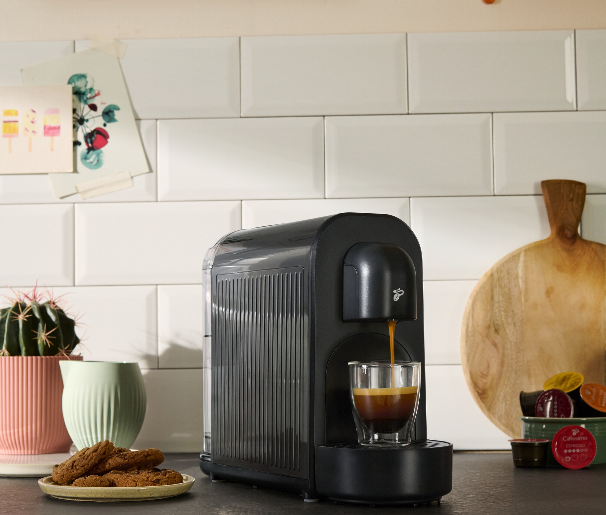 Čierny kávovar plní šálku kávou. Vidieť aj sušienky, sukulent a kuchynské dekorácie.