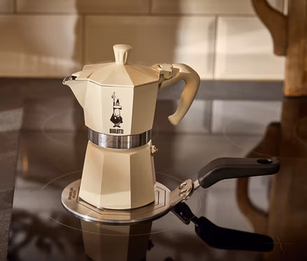 Krémový kávovar na espresso Bialetti Moka Express 190 ml stojí na adaptérovej podložke na indukciu Bialetti 13 cm vedľa pohára espressa.