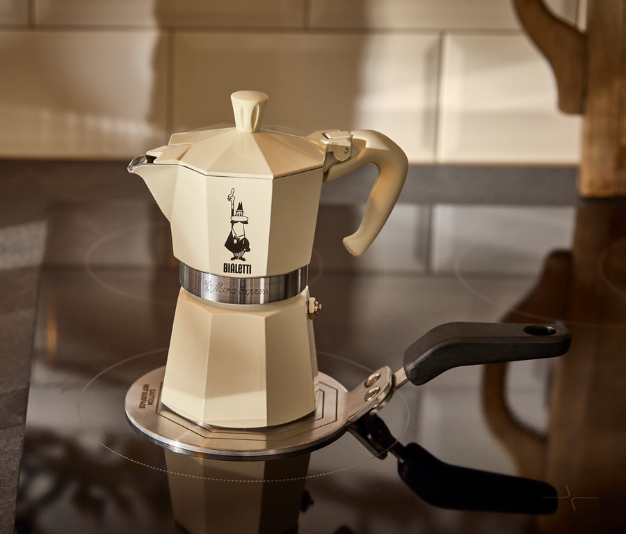 Krémový kávovar na espresso Bialetti Moka Express 190 ml stojí na adaptérovej podložke na indukciu Bialetti 13 cm vedľa pohára espressa.