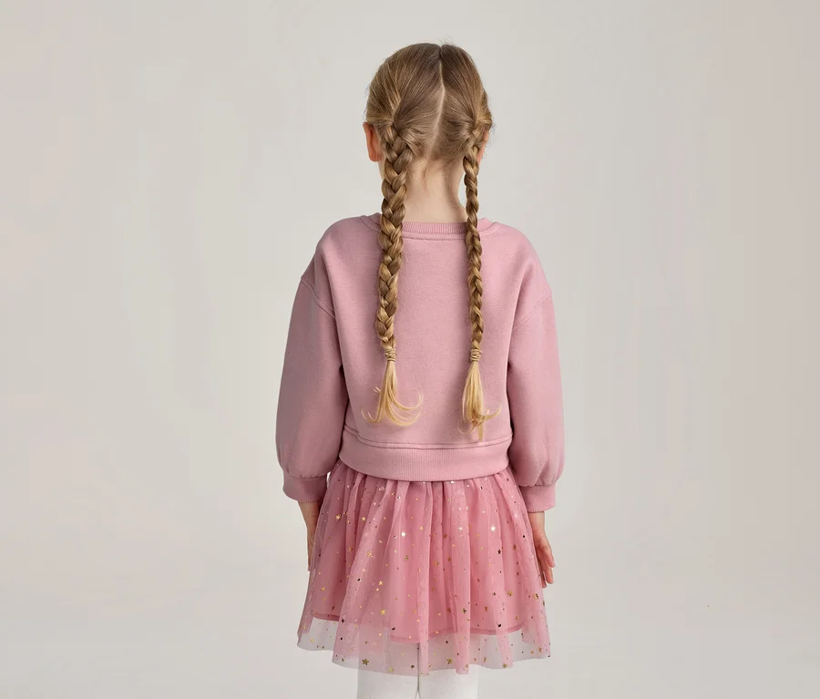 Dievča zozadu s blond vrkočmi, v ružovej mikine a ružovej tylovej sukni so zlatými hviezdičkami.
