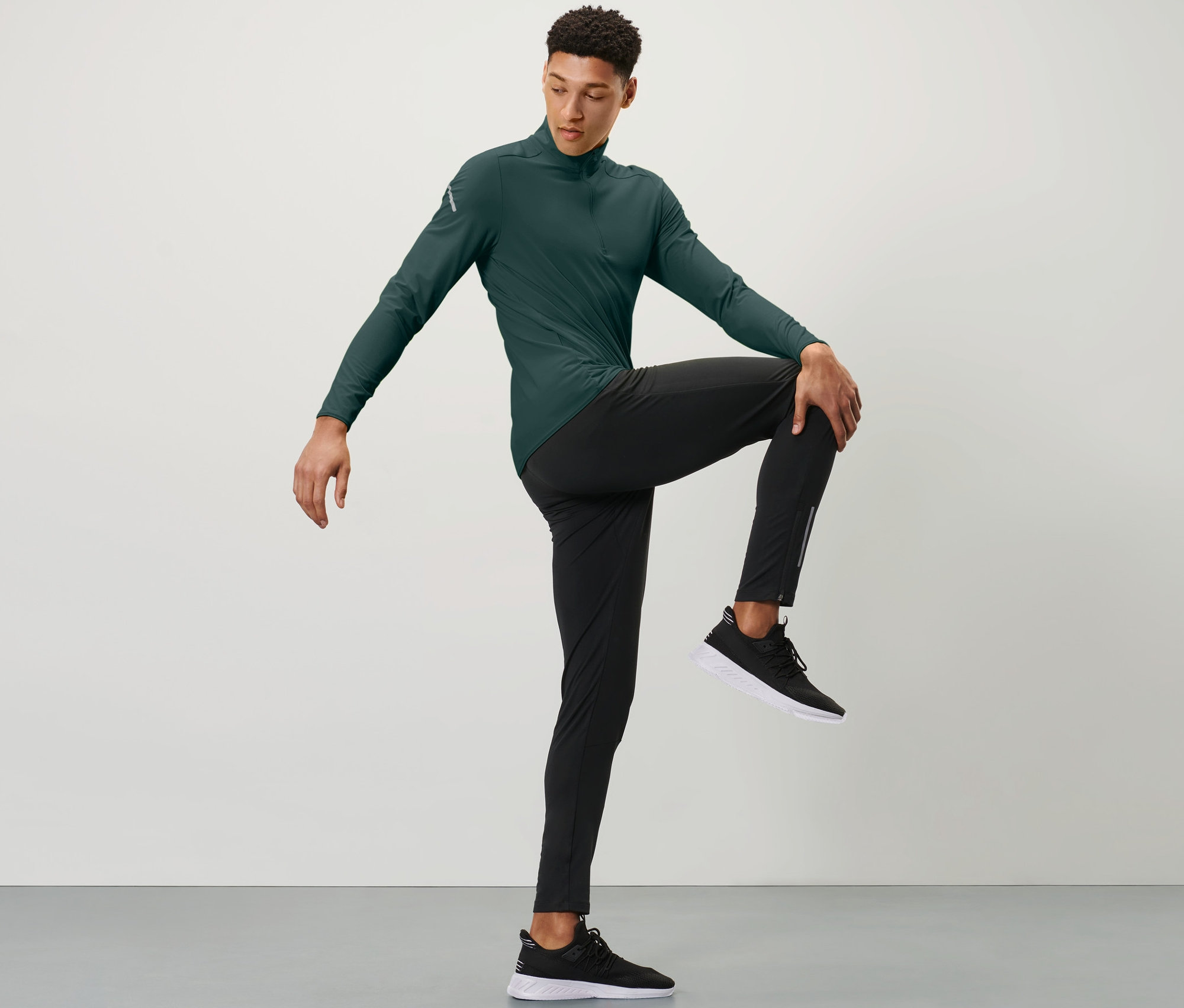Muž v zelenom športovom tričku a čiernych bežeckých legínach s ochranou proti vetru drží jedno koleno rukou pred hrudníkom.
