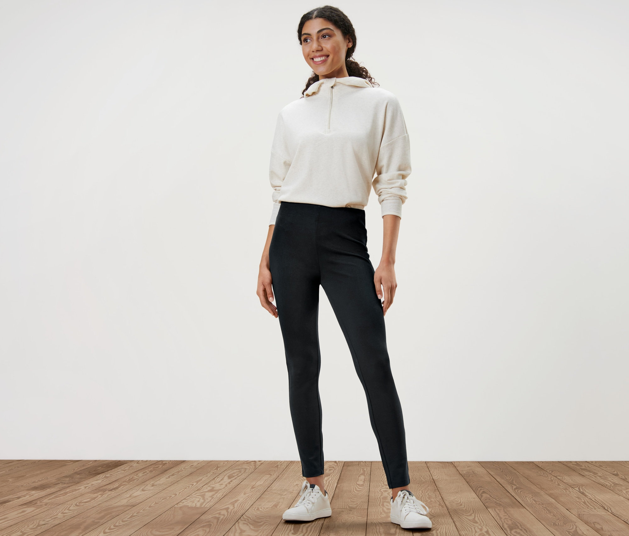 Žena stojí a má na sebe hrubé legínsy, bielu mikinu a biele tenisky.