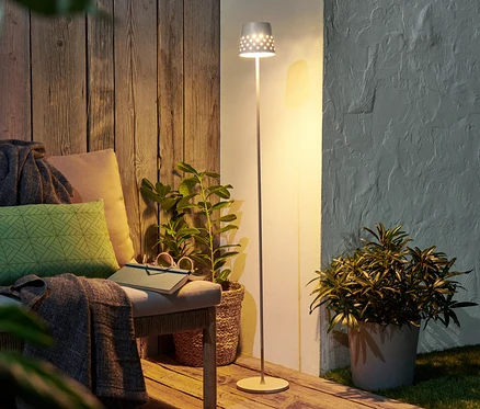 Kovová stojacia LED lampa na akumulátor stojí vedľa lavice s vankúšmi a knihou.