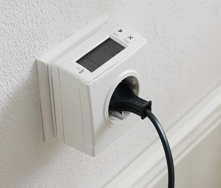 Na bielej stene je zásuvka s meračom spotreby elektrickej energie a zapojenou čiernou šnúrou.