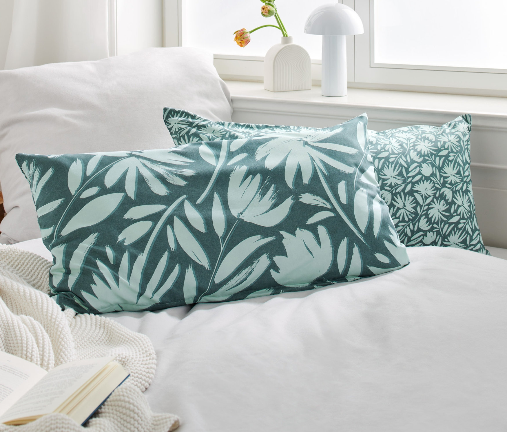 Dve zelené obliečky na vankúše z mikrovlákna s kvetinovým vzorom ležia na posteli vedľa otvorenej knihy.