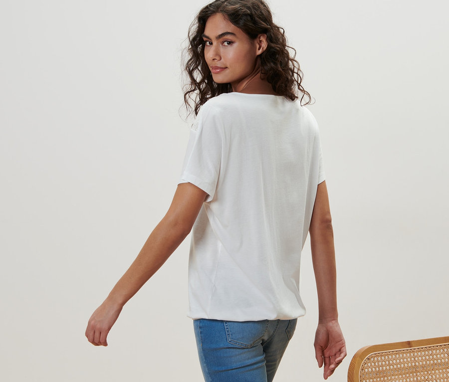 Žena kráča a pozerá sa cez plece. Má na sebe biele blúzkové tričko s výšivkou a džínsy.